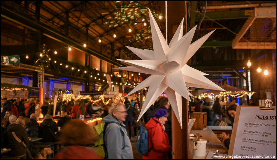 Ein weißer Stern an einem Weihnachtsmarkt-Stand. Dahinter unscharf Weihnachtsmarkt in einer Halle
