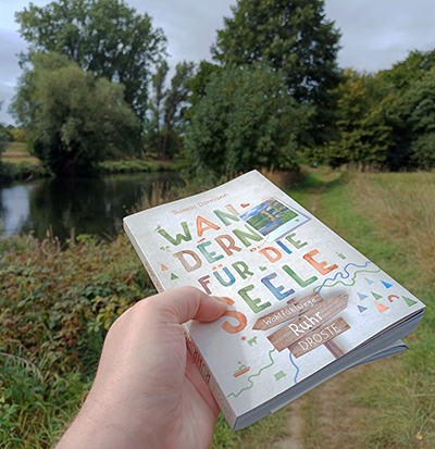 Eine Hand hält ein Buch "Wandern für die Seele" in der Hand vor einer Szene an einem Fluss.
