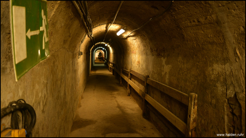 Langer Gang des Bunkers, bedeckt von meterdicken Massen von Abraum der Halde. Auf Holzbänken saßen die Menschen,  spürten und hörten die nahen Einschläge hautnah und hofften, dass es bald vorbei sein würde mit dem Bombardement. 