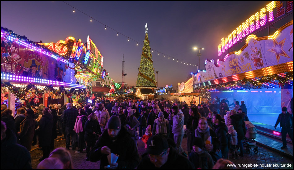 Kirmes-Stimmung mit Autoscooter und Buden und einem Weihnachtsbaum im Hintergrund. Zu sehen sind viele Menschen, die über die Kirmes laufen.