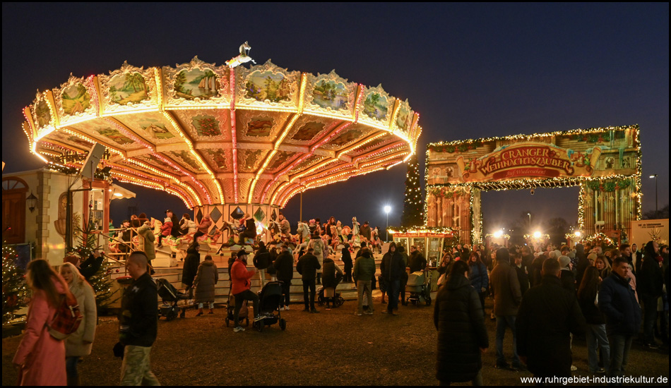 Ein klassisches Kinderkarussell mit Pferdchen in der Dunkelheit beleuchtet. Dahinter Eingang zum Cranger Weihnachtszauber