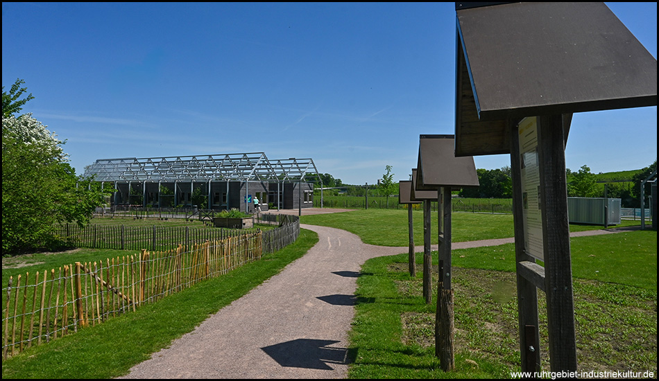 Blick auf einen Wanderweg mit Infotafeln, der zu dem modernen Gebäude mit Stahlkonstruktion und umzäuntem Garten führt.