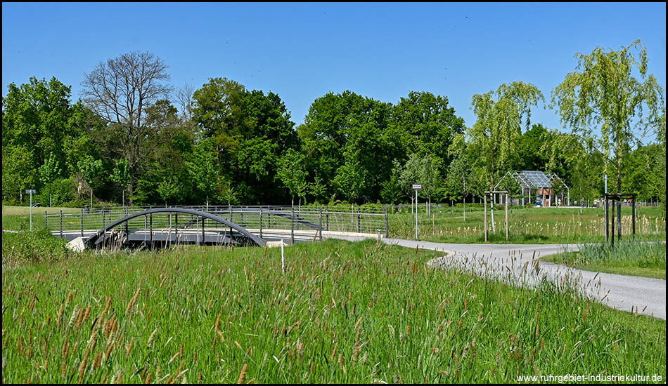 Wege führen durch eine Parklandschaft mit Wiesen und Bäumen. Eine Brücke mit einem charmanten Bogen führt über einen Bach
