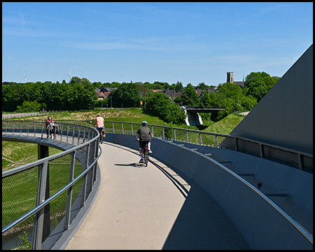 Radfahrer auf einem geschwungenen, erhöhten Radweg mit Metallgeländer, im Hintergrund eine grüne Landschaft mit einem Kirchturm