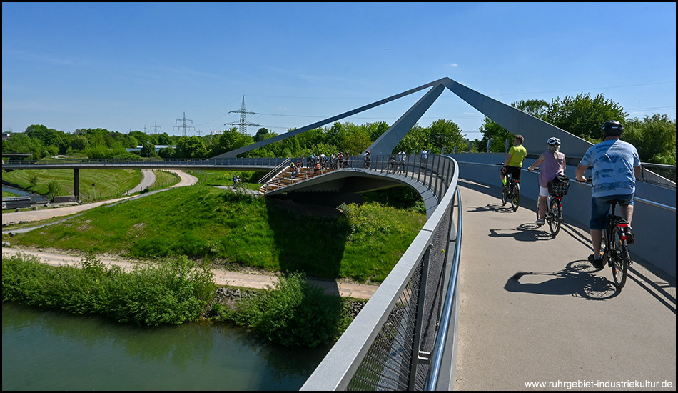 Radfahrer auf einer eleganten Brücke mit charakteristischer dreieckiger Stahlkonstruktion über einem kleinen Fluss und grünen Wiesen.
