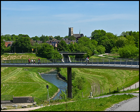 Eine moderne Fußgänger- und Radfahrerbrücke über einen Fluss, mit Blick auf ein Dorf und eine Kirche im Hintergrund.