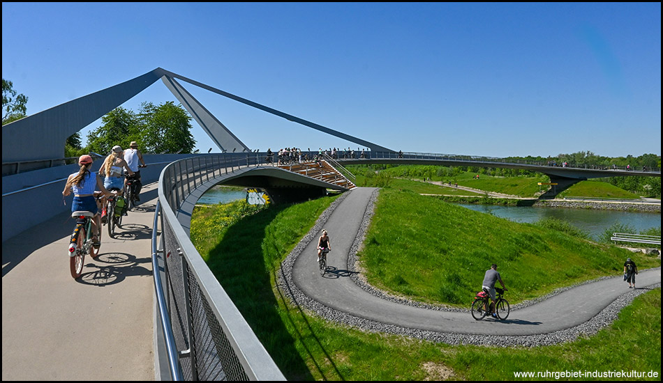 Radfahrer auf einer spektakulären Brücke mit großer Stahlkonstruktion, darunter gewundene Radwege durch die grüne Parklandschaft.