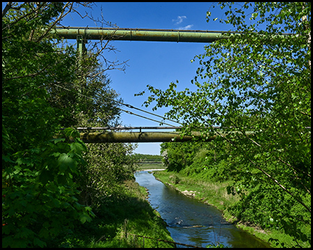 Zwei industrielle Rohrleitungen überqueren einen schmalen Fluss, der von grünen Bäumen und Büschen umgeben ist. Das klare Wasser fließt ruhig unter den Rohren hindurch bei blauem Himmel.