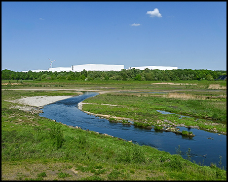 Ein mäandernder blauer Wasserlauf schlängelt sich durch eine offene Landschaft mit niedrigem Bewuchs. Im Hintergrund steht ein großes weißes Industriegebäude, umgeben von grünen Bäumen unter einem klaren Himmel.
