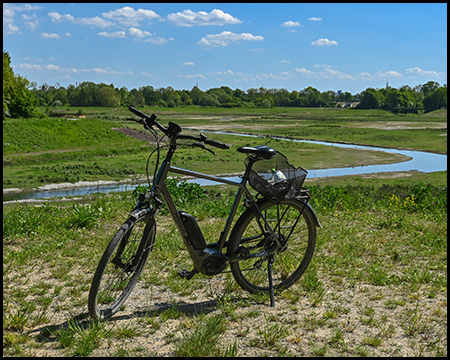 Ein schwarzes Elektrofahrrad steht auf einer leicht erhöhten Position mit Blick auf eine Auenlandschaft. Ein schmaler Fluss schlängelt sich durch die grüne Fläche unter blauem Himmel mit weißen Wolken.
