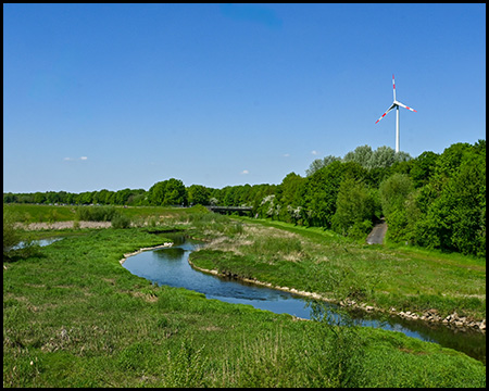 Ein schmaler blauer Fluss schlängelt sich durch eine grüne Landschaft. Am rechten Bildrand stehen dichte Bäume, im Hintergrund ist eine Windkraftanlage zu sehen. Der Himmel ist strahlend blau mit einigen kleinen weißen Wolken.