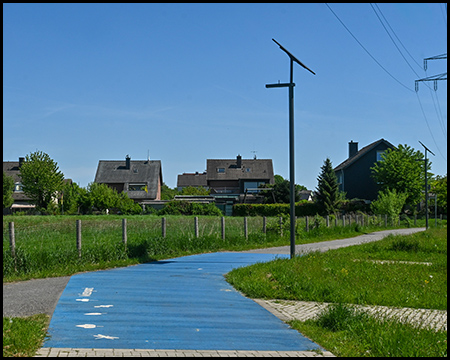 Ein blau markierter Radweg verläuft durch eine grüne Landschaft. Am Wegrand steht eine von einer Solarzelle gespeiste Laterne. Im Hintergrund sind mehrere Einfamilienhäuser zu sehen. Der Himmel ist klar und blau.