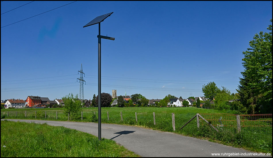 Eine sonnige Landschaftsaufnahme mit einem Feldweg am Rand einer grünen Wiese. Im Vordergrund steht eine Straßenlaterne mit Solarpanel, im Hintergrund sind ein Dorf mit Kirchturm und Stromleitungen zu sehen.