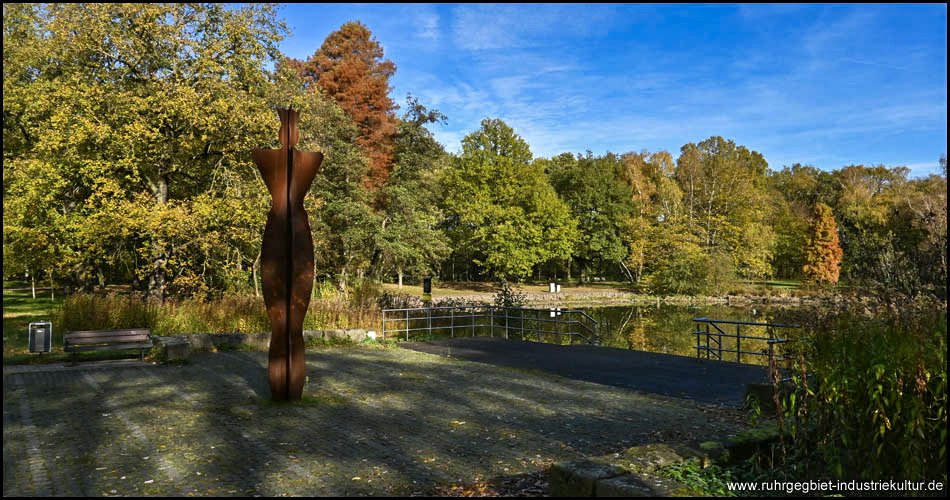 Teich mit im Wasser spiegelnden Uferbäumen, Terrasse und Skulptur