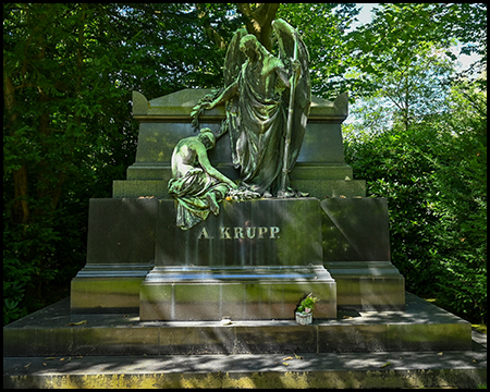 Monumentales Krupp-Familiengrabmal mit dramatischen Bronzeskulpturen. Eine große, geflügelte Figur steht aufrecht, während eine zweite Figur sitzend dargestellt ist. Der massive Steinsockel trägt den Namen "A. KRUPP" in goldenen Buchstaben. Das Grabmal ist von grünen Bäumen umrahmt und steht auf einer mehrstufigen Plattform aus hellem Stein.