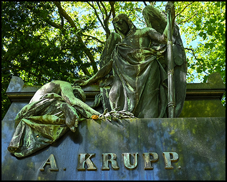 Nahaufnahme der kunstvollen Bronzeskulpturen auf dem Krupp-Grabdenkmal. Eine geflügelte Figur mit Stab steht erhaben neben einer sitzenden, trauernden Gestalt, die einen Kranz hält. Darunter ist der Name "A. KRUPP" in großen, goldenen Buchstaben auf dem dunklen Steinsockel zu sehen.