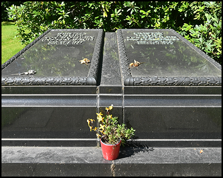 Doppelgrabstätte mit zwei nebeneinanderliegenden dunklen Steinplatten, beide umrahmt von einem Ornamentband. In der Mitte zwischen den Platten steht ein kleiner roter Blumentopf mit gelben Blüten. Auf den Platten sind die Namen und Lebensdaten der Verstorbenen eingraviert.