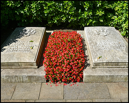 Doppelgrabstätte mit zwei steinernen Grabplatten, die durch einen üppigen Streifen von leuchtend roten Blumen verbunden sind. Die steingrauen Tafeln tragen reliefartige Verzierungen an ihren oberen Rändern und Inschriften mit Namen und Daten der Verstorbenen. Das Grab ist auf einer gepflasterten Fläche angelegt und wird von grünen Sträuchern eingerahmt.
