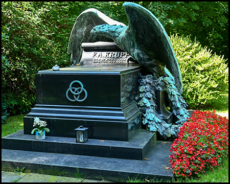 Nahaufnahme des markanten Krupp-Grabmals mit dem mächtigen Bronzeadler, dessen ausgebreitete Flügel schützend über dem schwarzen Sarkophag ruhen. Auf der Vorderseite des Sarkophags ist "F.A. KRUPP" und die Lebensdaten zu lesen, sowie ein kreisförmiges Symbol mit drei ineinander verschlungenen Ringen. Am Fuße des Denkmals stehen kleine Laternen und ein prächtiges Beet mit leuchtend roten Blumen schmückt die rechte Seite.