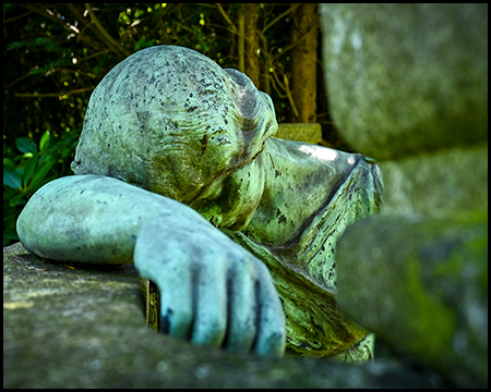 Verwitterte Steinskulptur eines trauernden Wesens. Die vom Alter gezeichnete Figur mit sanften Konturen scheint auf einer Steinoberfläche zu ruhen. Das moosige Grün der Patina verschmilzt mit der umgebenden Vegetation und vermittelt Zeitlosigkeit und Vergänglichkeit.