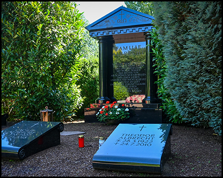Aufwändiges Familiengrab mit klassizistischem Tempelmotiv in Blau und Schwarz. Die Grabstätte ist von hohen Hecken umgeben und mit Blumen geschmückt.