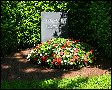 Schlichter Grabstein für Karl und Mia Albrecht, umrahmt von üppigem Grün und geschmückt mit einem farbenfrohen Blumenbeet aus roten und weißen Blüten.