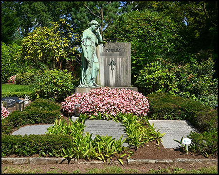 Kunstvoll gestaltetes Grab mit stehender Bronzefigur neben einer Steinplatte mit Kreuzemblem. Das Monument ist von einem Meer aus rosa Blumen umgeben und in eine strukturierte Gartenanlage eingebettet
