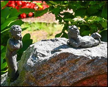 Detailaufnahme kleiner Bronzefiguren auf einem Naturstein. Die filigranen Skulpturen stellen kindliche Gestalten dar, die auf dem rauen Gestein zu spielen oder zu ruhen scheinen.