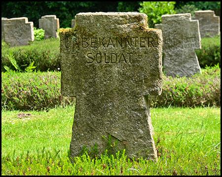 Ein verwitterter Grabstein trägt die eingemeißelte Inschrift "UNBEKANNTER SOLDAT". Das schlichte Steinkreuz steht zwischen anderen Gräbern auf grünem Rasen