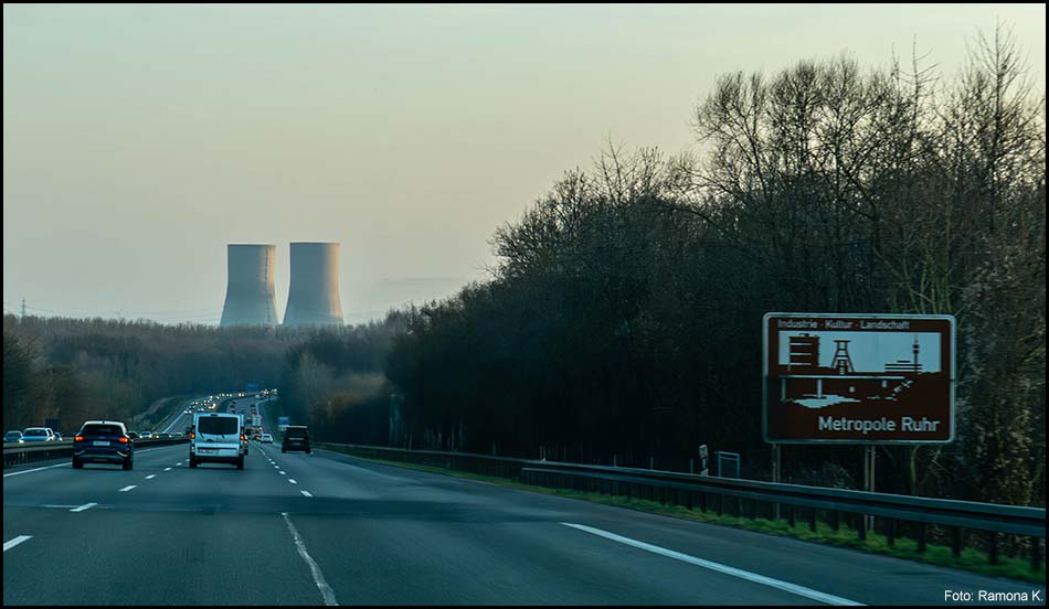 Ansicht aus einem Auto auf der Autobahn. Vorne steht eine Unterrichtungstafel, die den Beginn der Metropole Ruhr anzeigt. Am Horizont ist ein Kraftwerk zu sehen.