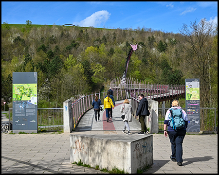 ußgängerbrücke mit pinken Elementen und einer drachenartigen Skulptur am Ende. Besucher überqueren die Brücke vor einem bewaldeten Hügel