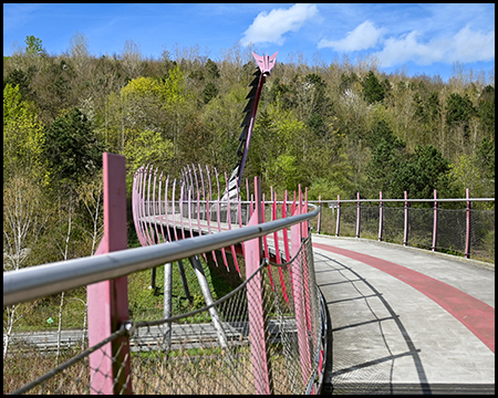 Nahaufnahme der geschwungenen pinken Brücke mit einem Drachenkopf am Ende, der sich über einen bewaldeten Hintergrund erhebt.