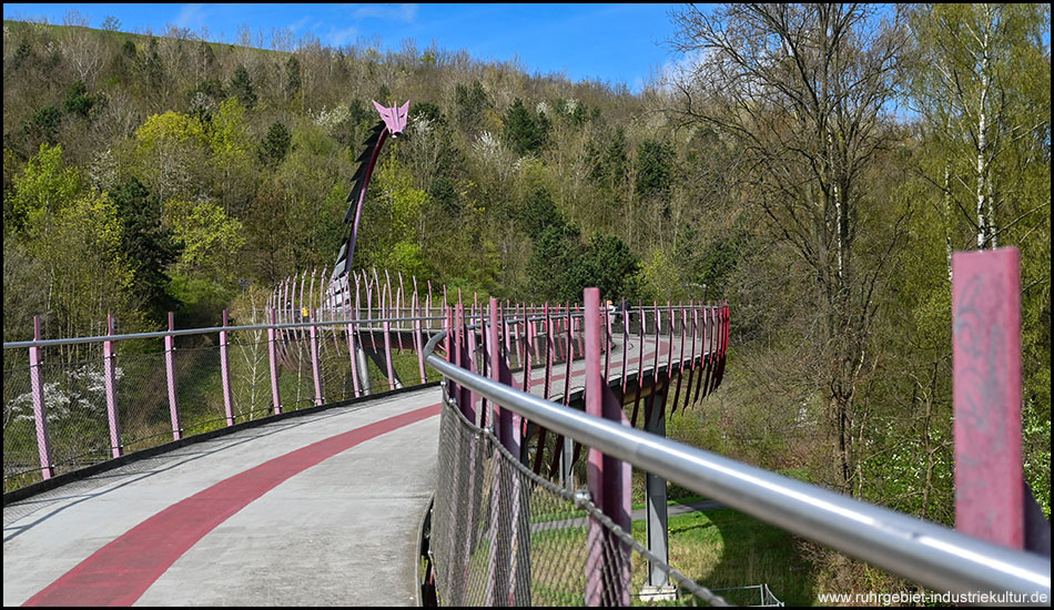 Perspektive entlang der geschwungenen Brücke mit pinken Stützen und einem Drachenkopf, umgeben von Frühlingsbäumen am Hang.