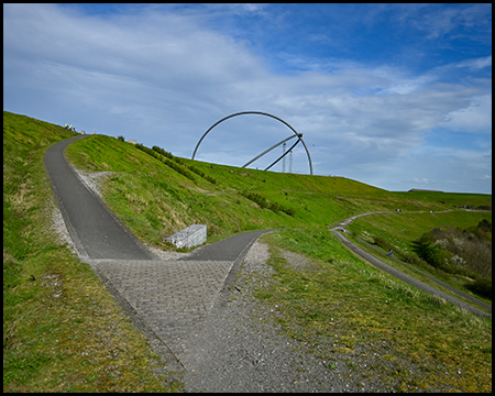 Gepflasterte Wege am grünen Hügel mit einer großen bogenförmigen Metallskulptur auf dem Gipfel.