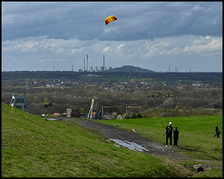 Menschen beim Drachenfliegen auf einer Hügelkuppe mit Blick auf eine Siedlung und Industrieanlagen in der Ferne.