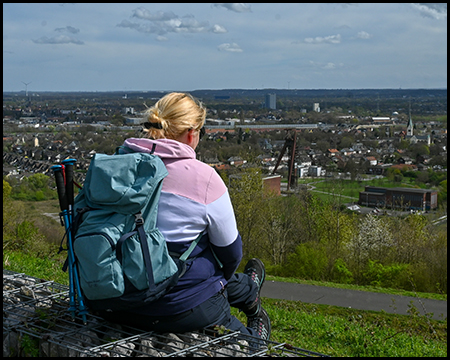Eine Person mit Rucksack sitzt auf einem Gabionen-Block und blickt über eine städtische Landschaft.