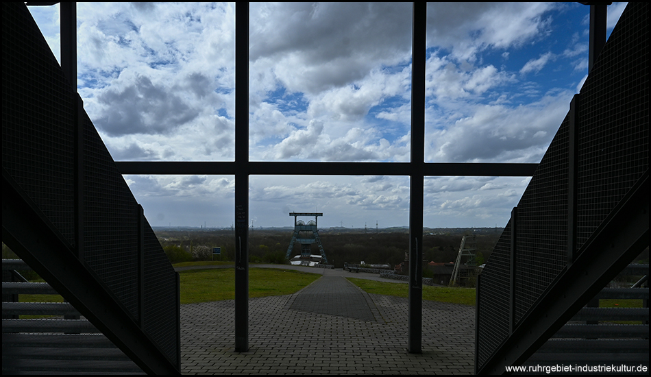Blick durch ein architektonisches Fenster auf einen Förderturm und die umliegende Landschaft.