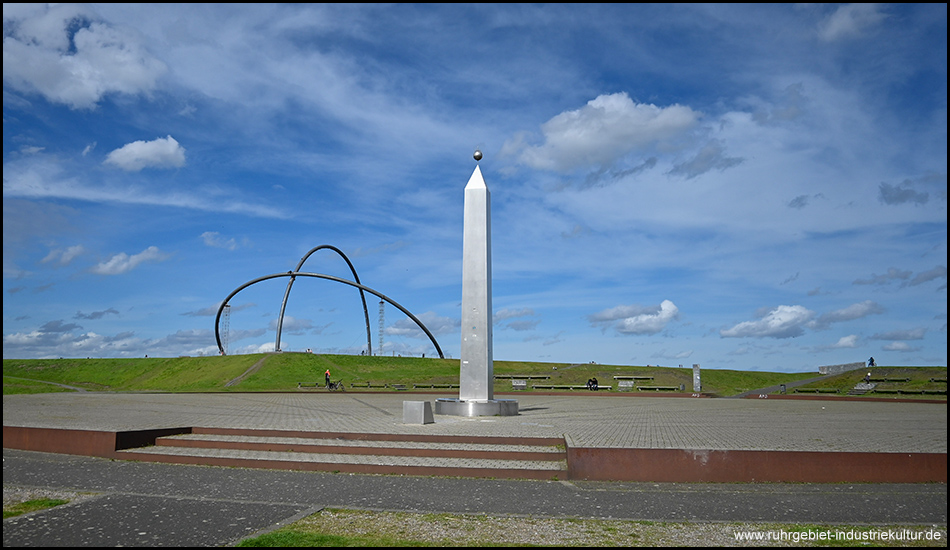 Obelisk im Vordergrund mit der stählernen Bogenstruktur auf dem grünen Hügel im Hintergrund.
