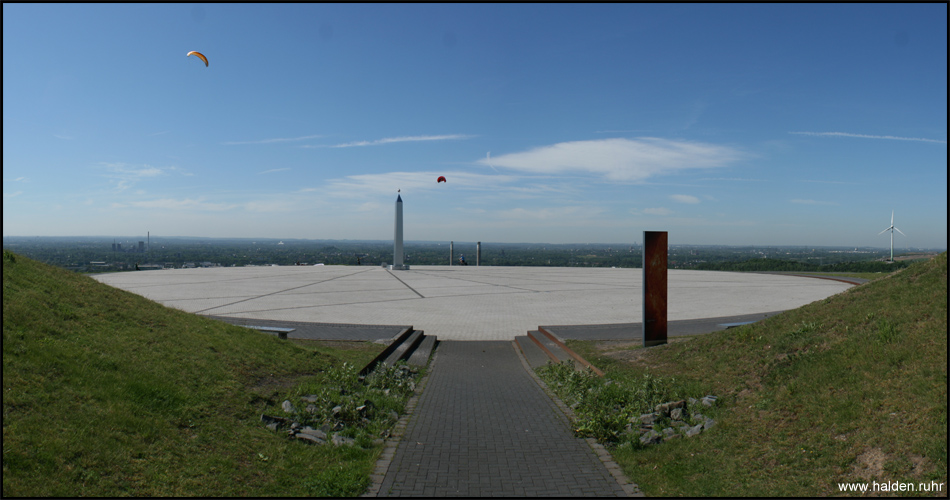 Panoramablick auf das kreisrunde Plateau mit der großen Sonnenuhr auf der Halde Hoheward, rechts Windrad von Halde Hoppenbruch