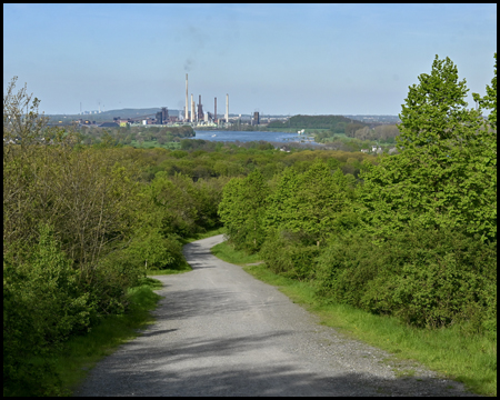 Weg auf einer Halde bergab in eine Ebene mit einem Fluss. Im Hintergrund Industrie