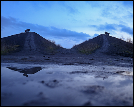 Zwei dunkle Hügel mit technischen Installationen auf den Spitzen, im Vordergrund eine Pfütze, die den blauen Abendhimmel spiegelt.