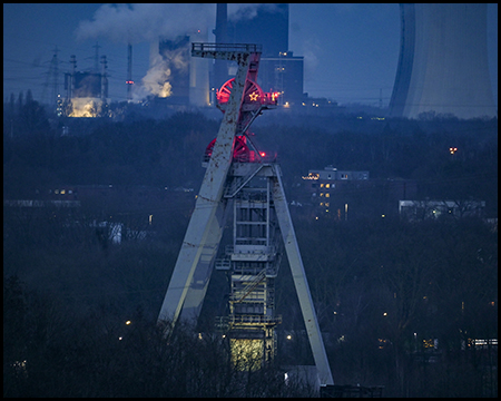 Ein beleuchteter Förderturm mit roten Lichtern vor einem dunklen Himmel, im Hintergrund Fabrikgebäude mit Dampfausstoß.
