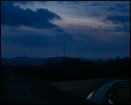 Eine einsame Windkraftanlage am Horizont vor einem dunkelblauen Abendhimmel, im Vordergrund dunkle Landschaft.