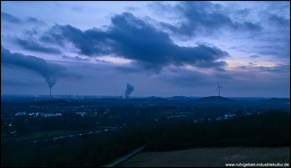 Panoramablick über eine Industrielandschaft bei Nacht mit Fabrikschornsteinen, Windrädern und städtischen Lichtern.