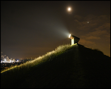 Ein beleuchteter Scheinwerfer auf einem Hügel bei Nacht, der Mond und Stadtlichter sind am Horizont sichtbar.