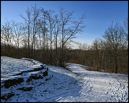 Verschneiter Hangweg mit Spurrillen, gesäumt von kahlen Bäumen und Felsen, unter teilweise bewölktem Himmel.