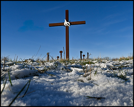 Stahlkreuz mit Tauben-Symbol auf verschneiter Anhöhe, im Hintergrund weitere Pfosten unter blauem Himmel, Froschperspektive.