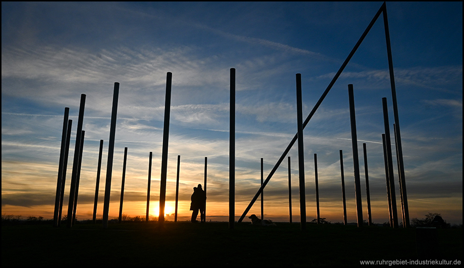 Sonnenuhr auf der Halde Schwerin im Gegenlicht des Sonnenuntergangs mit einem Pärchen und einem Hund als schwarze Silhouette