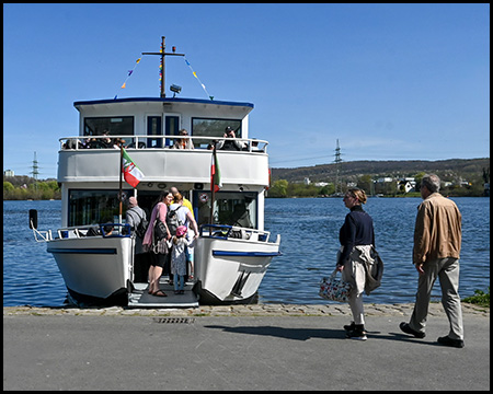 Das Ausflugsschiff liegt am Ufer an. Passagiere steigen über die vordere Rampe ein oder aus. Zwei Personen stehen am Ufer und warten. Am Oberdeck des Schiffes sind NRW-Flaggen zu sehen. Die Umgebung zeigt eine Seenlandschaft mit Hügeln im Hintergrund.