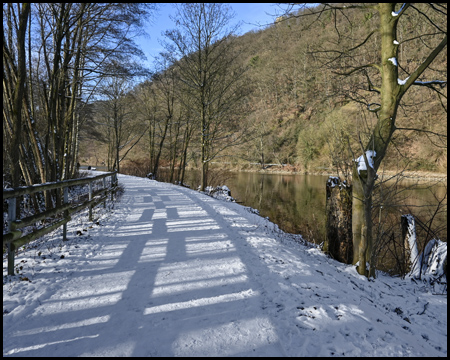 Ein Weg am Wasser entlang mit Schnee und Schatten von einem Zaun daneben